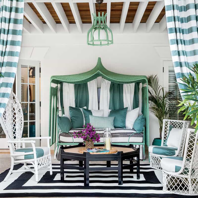 downstairs patio, teal and white striped curtains, 3 white chairs, black coffee table with purple flowers and a candle on a black and white rug, teal light fixture above, teal and while cabana with teal and white striped pillows,