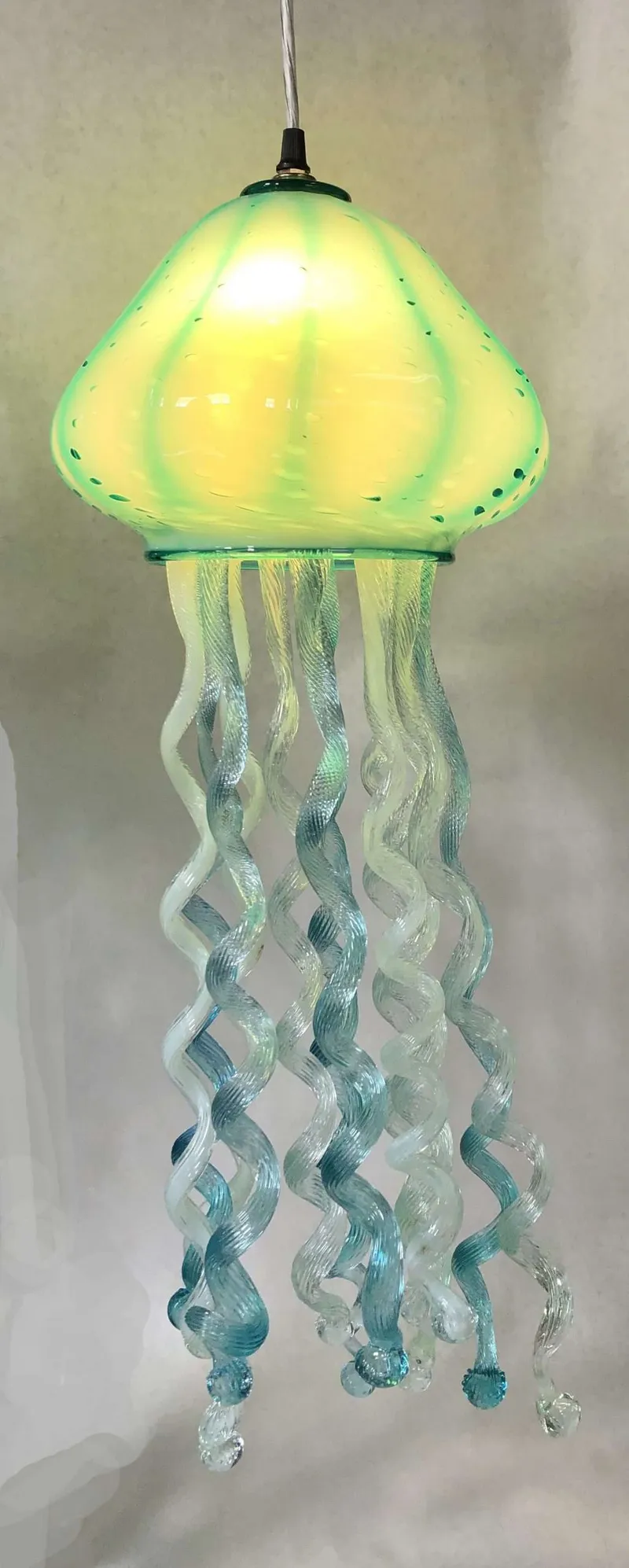 This vibrant blown glass pendant light mimics a jellyfish, featuring a glowing chartreuse bell with textured blue-green tentacles, creating unique modern interior lighting.