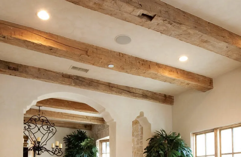 Rough-hewn faux reclaimed wood ceiling beams with a natural texture define this rustic interior's open-concept design featuring smooth plastered walls and arched architectural elements.