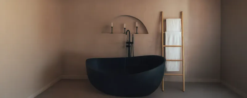 A contemporary bathroom features a matte black freestanding tub, textured clay-based walls with an arched niche, and a natural wooden ladder, creating a serene, modern interior design.