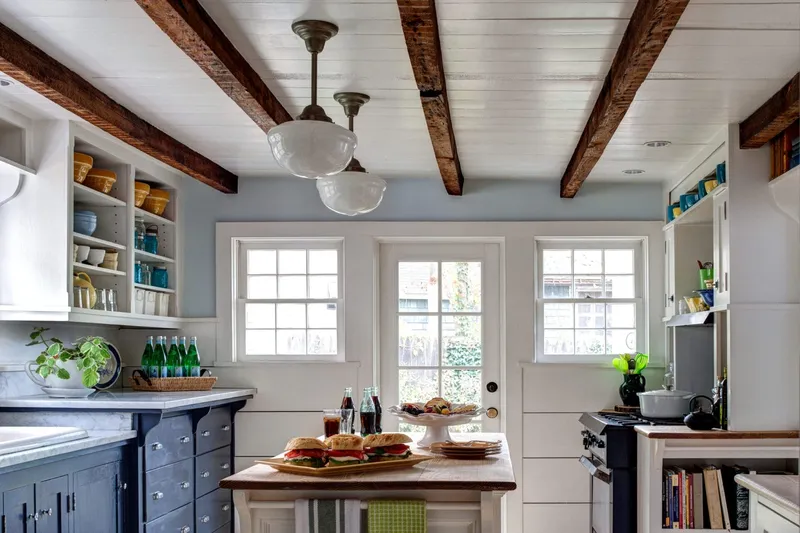 A charming kitchen features rough-hewn faux reclaimed wood beams contrasting with white plank ceilings, blue cabinetry, and clean shaker-style shelving for a rustic modern design.