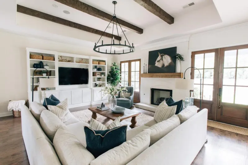 A welcoming living room features a vaulted ceiling with faux reclaimed wood beams, complemented by a large sectional, plush rug, and rustic wooden coffee table.