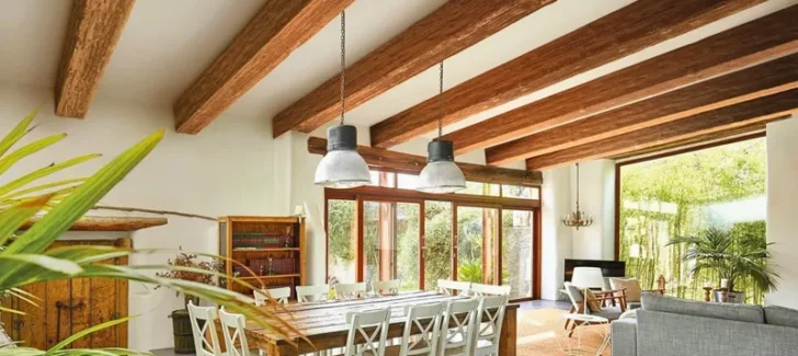 This open-plan interior design features a dining area with a rustic wood table and white chairs, overlooked by impressive faux reclaimed wood ceiling beams and large windows, connecting to a cozy living space.