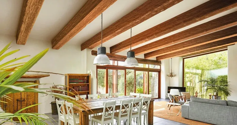 This open-plan interior design features a dining area with a rustic wood table and white chairs, overlooked by impressive faux reclaimed wood ceiling beams and large windows, connecting to a cozy living space.