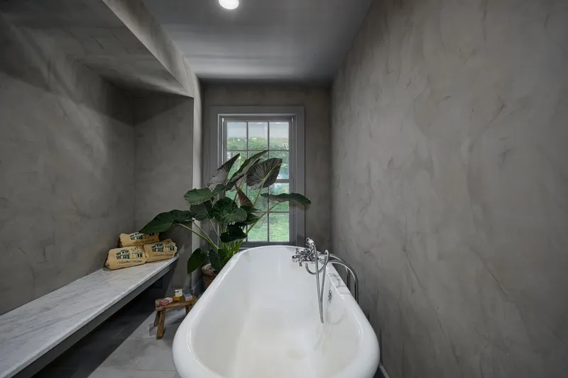 A tranquil modern bathroom features textured clay-based walls and a pristine white freestanding tub, complemented by a sleek marble bench and lush tropical plant, enhancing the interior design.