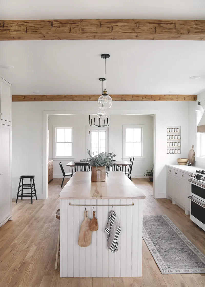 A modern farmhouse kitchen features rustic faux wood ceiling beams, warm light wood flooring, a white beaded kitchen island, and contemporary lighting, creating an inviting open-concept design.