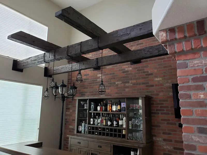 Dramatic faux reclaimed wood ceiling beams with a deeply textured finish add rustic charm and a grand architectural element above a custom home bar and exposed brick wall.