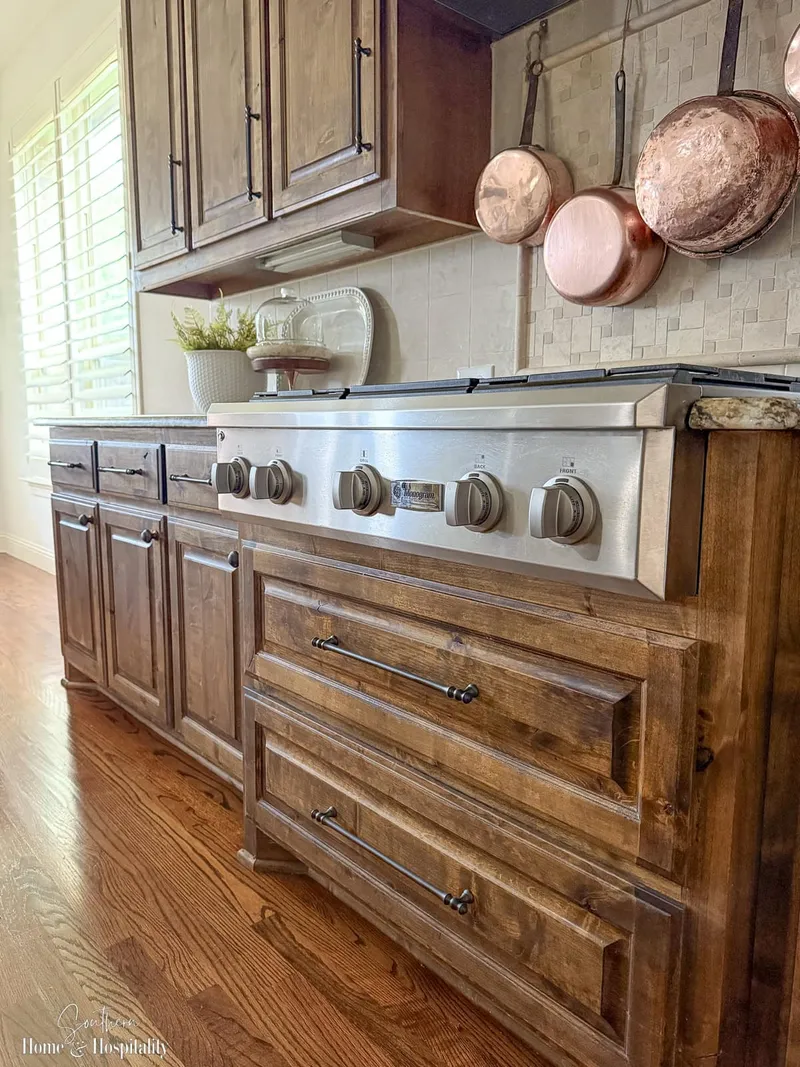 A warm, modern kitchen features rich stained wood cabinet fronts with traditional paneling, sleek stainless steel appliances, a tiled backsplash, and gleaming copper pots.
