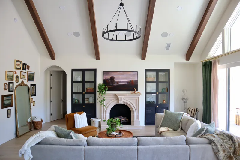 A bright living room showcases a vaulted ceiling with faux wood beams, an ornate stone fireplace, and custom black built-ins for a sophisticated interior design.