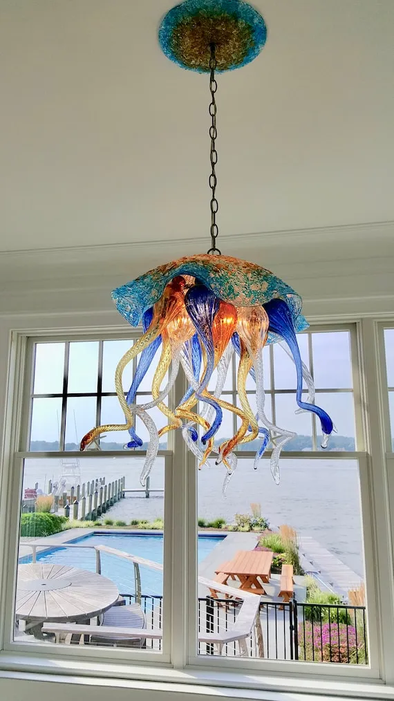 A vibrant glass jellyfish chandelier with textured blue and gold accents illuminates a bright interior, offering stunning waterside views of a pool deck and wooden pier.