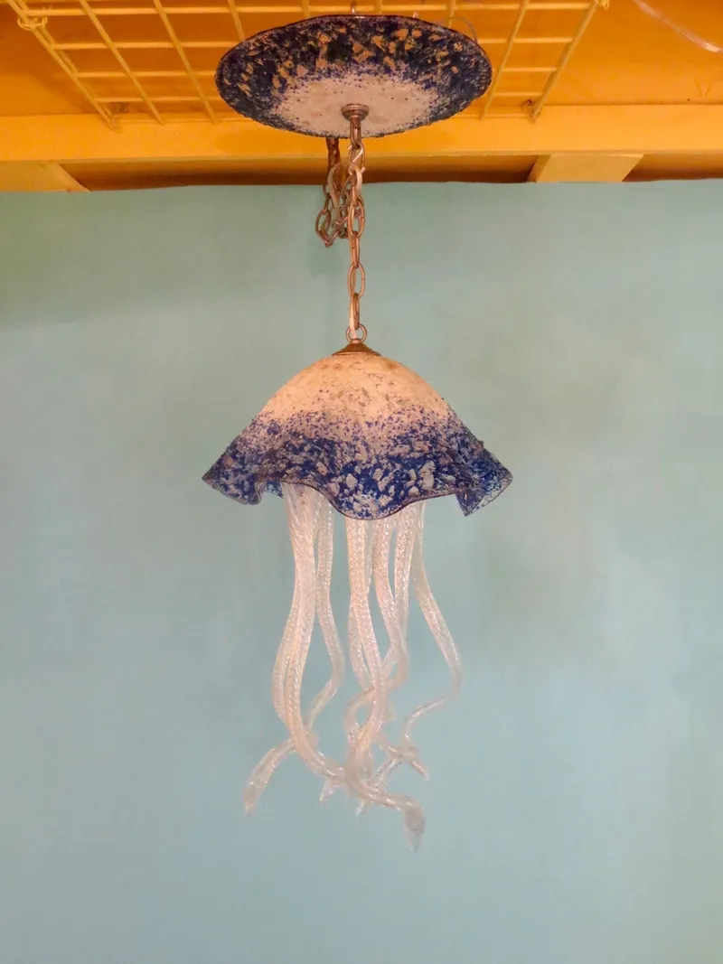 This distinctive jellyfish-inspired glass chandelier, featuring textured blue and white speckled shades and clear wavy tentacles, provides a unique lighting fixture against a pale blue wall.