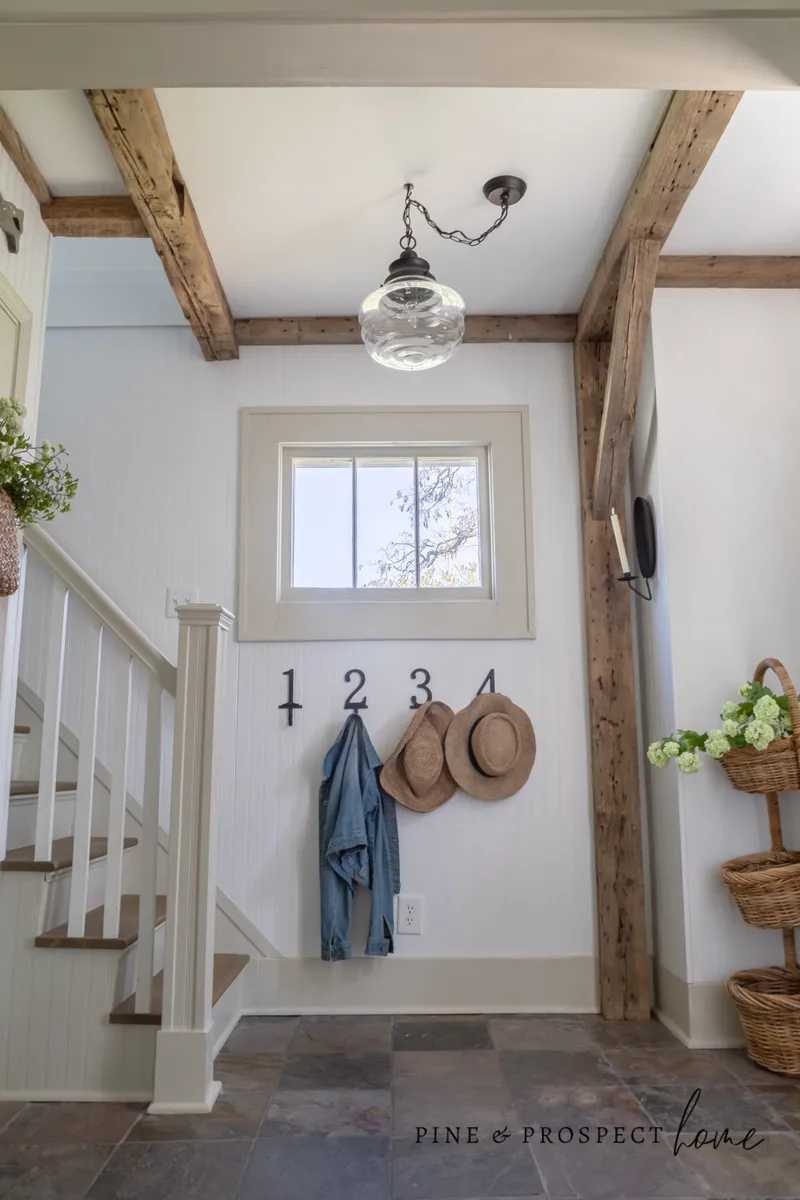 Rustic ceiling beams and a large clear glass pendant light illuminate this charming entryway featuring white beadboard walls, a staircase, and earthy slate tile flooring.