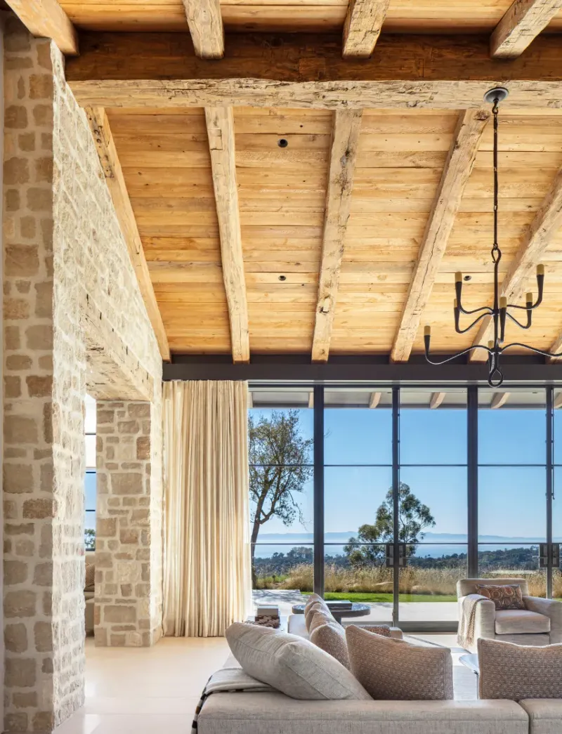 This rustic-modern interior design features stunning faux reclaimed wood ceiling beams, exposed stone walls, and expansive glass doors opening to a scenic landscape.