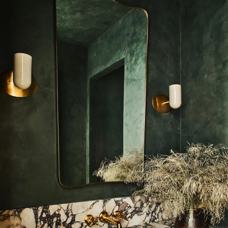 A luxurious modern bathroom features deep green clay-based wall finishes, a sleek brass-framed mirror, and a richly veined marble vanity with elegant brass fixtures.