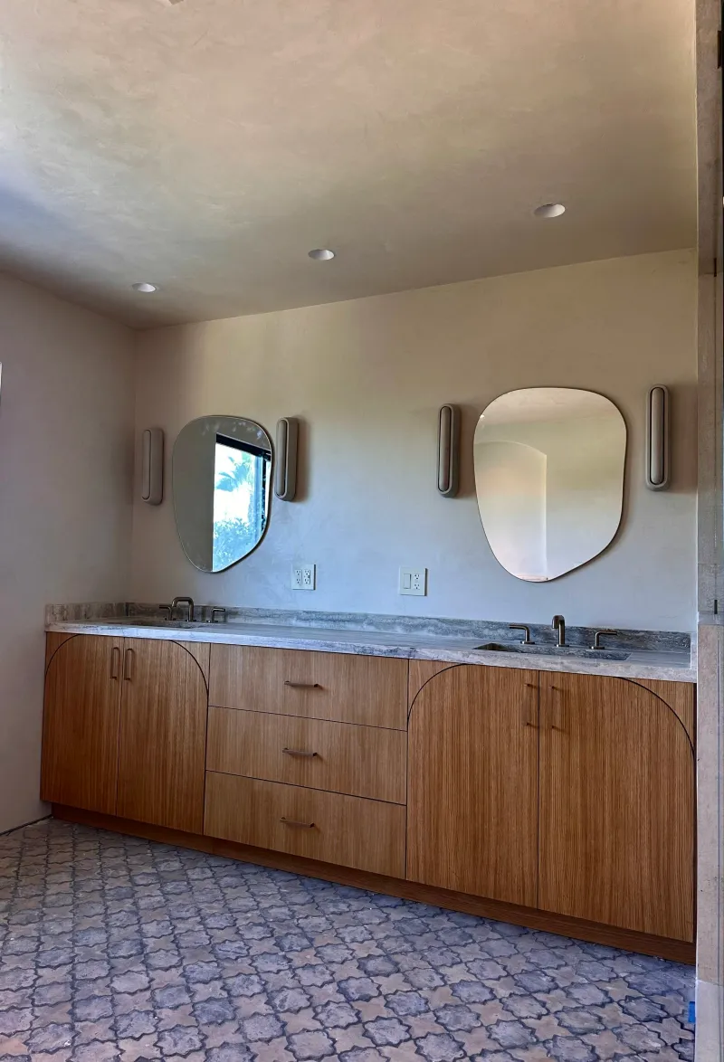 A modern bathroom vanity features warm wood cabinetry with curved details, a veined marble countertop, and organic mirrors against a softly textured clay-based wall finish.