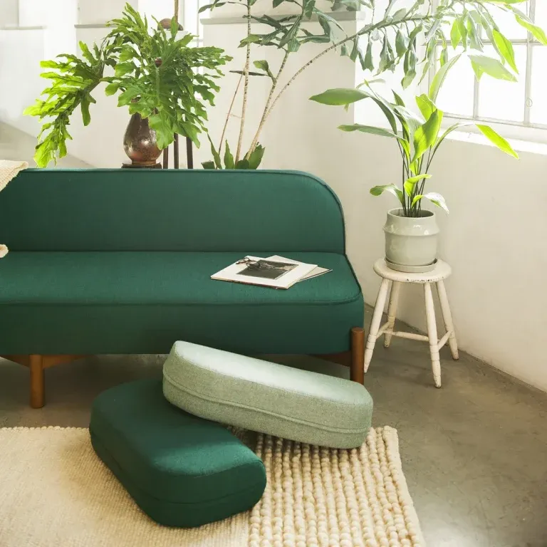 A modern interior features a sleek green upholstered sofa with curved lines, complemented by a textured natural fiber rug, soft ottomans, and lush potted plants.