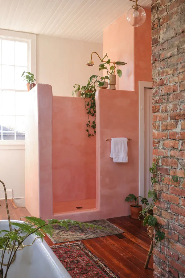 A beautifully textured clay-based shower enclosure in a warm blush hue creates a tranquil bathroom design, complemented by exposed brick and rich hardwood flooring.
