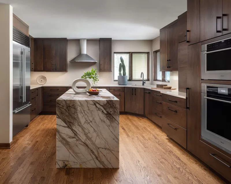 This modern kitchen design features warm, stained wood cabinetry, a striking marbled waterfall island, and rich hardwood flooring, creating a sophisticated and inviting interior.