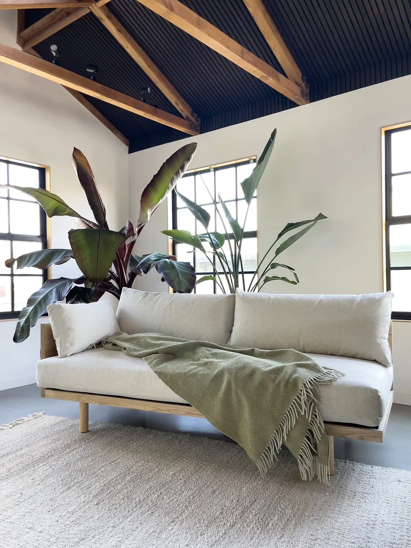 A contemporary living space features a light-toned natural fiber sofa with a textured green throw, complemented by a woven rug and exposed wooden ceiling beams.